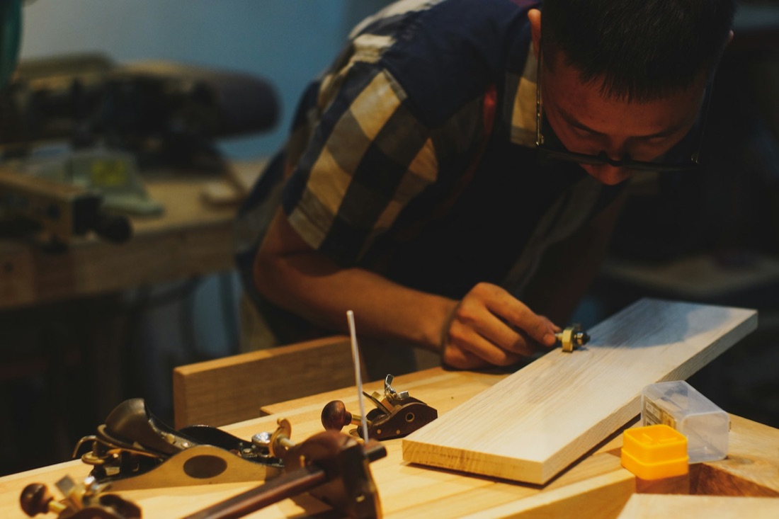 A carpenter marking a timber plank at a workbench in a warmly lit workshop